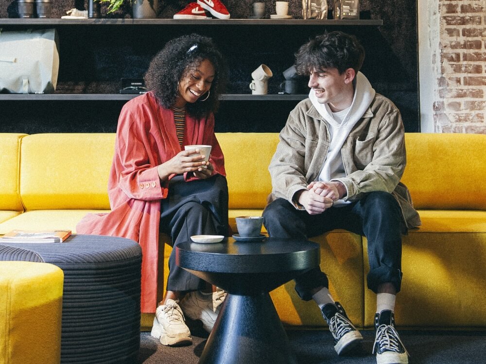 An image of a young woman and man sitting on a yellow sofa drinking coffee and smiling. 