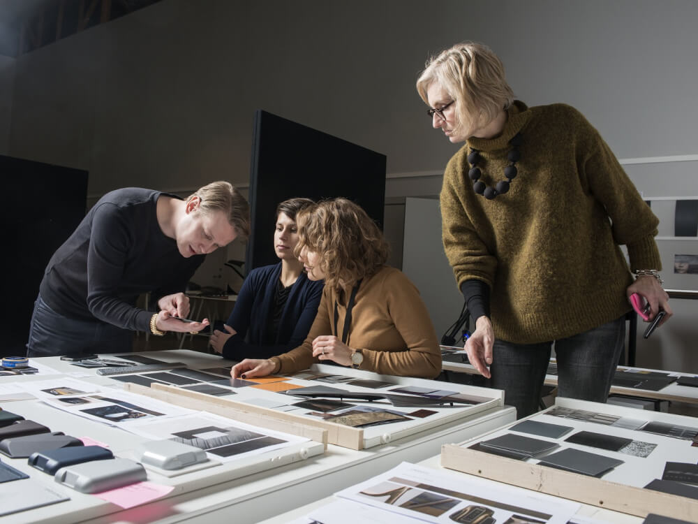 An image of four designers leaning over a table of fabric samples. One is holding a piece of fabric in his hand while the others look at what he is demonstrating. 