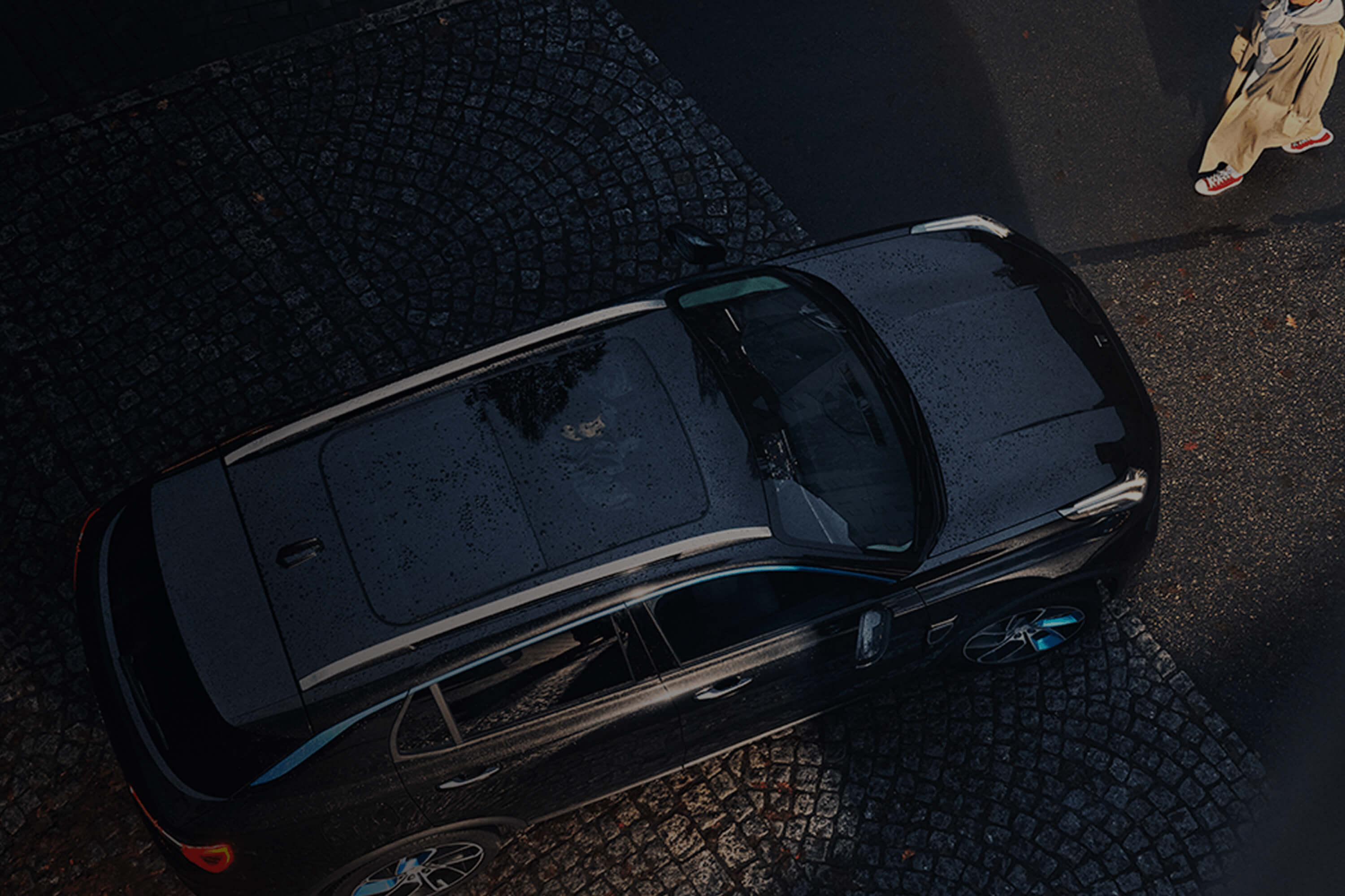 A top-down image of a woman approaching a Lynk & co car parked on a cobblestone street. A male driver is visible inside the car through the sunroof.