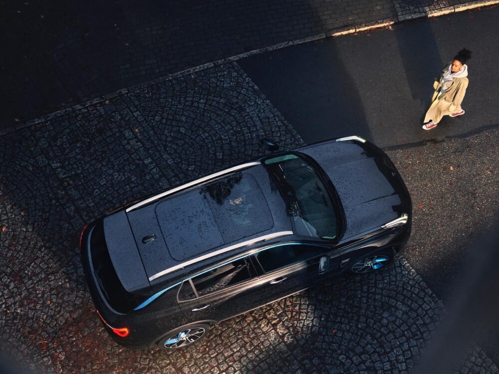 A top-down image of a woman approaching a Lynk & co car parked on a cobblestone street. A male driver is visible inside the car through the sunroof.